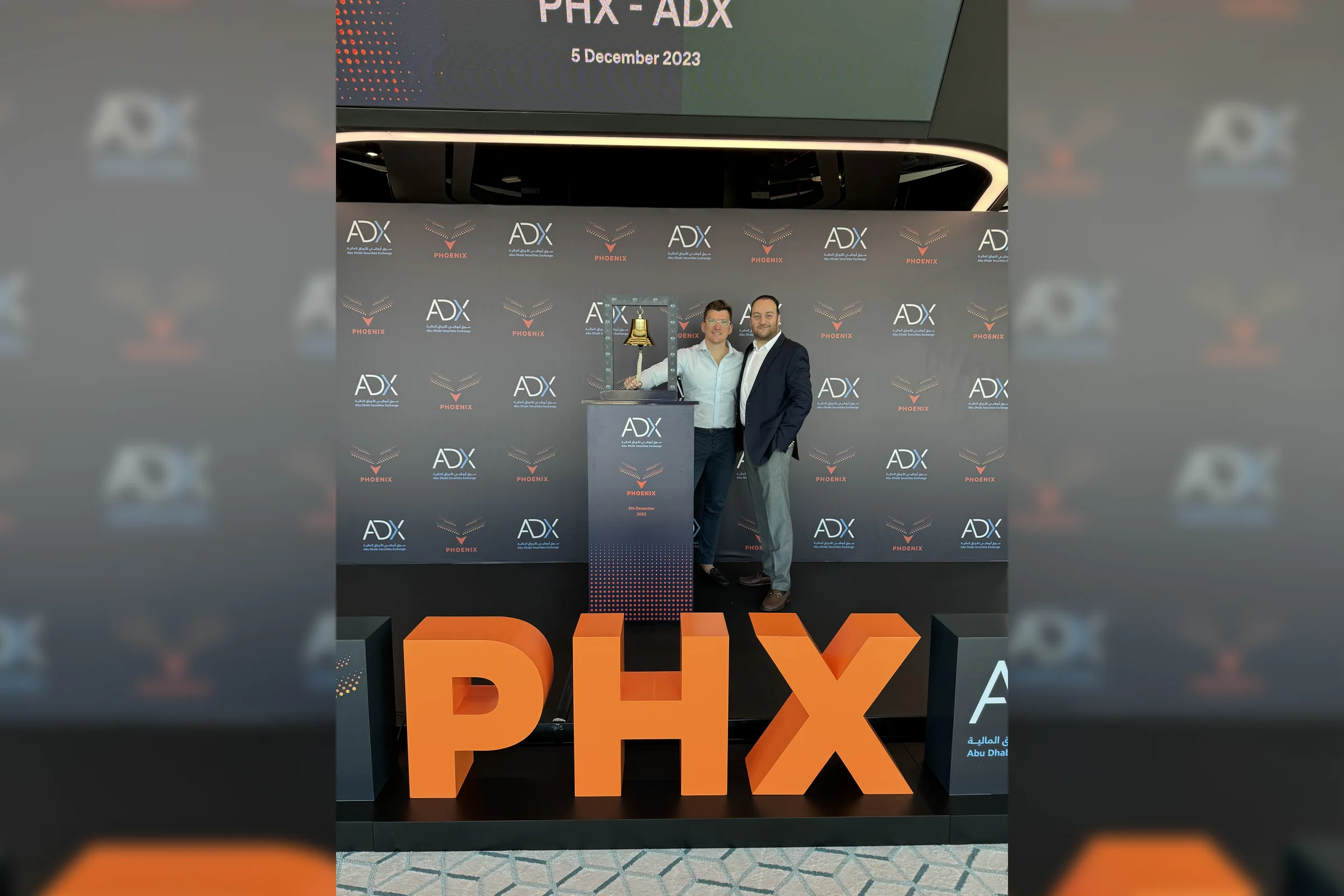 Two Phoenix Group colleagues beside the ceremonial bell and the PHX letters at the Abu Dhabi Securities Exchange