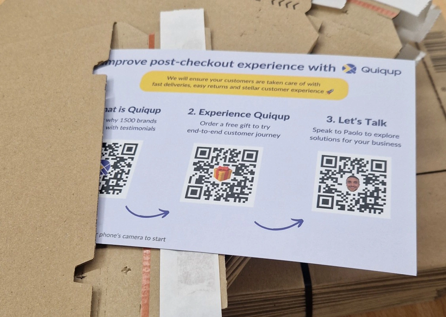 The printed Quiqup outreach card resting on a stack of flat-packed shipping boxes